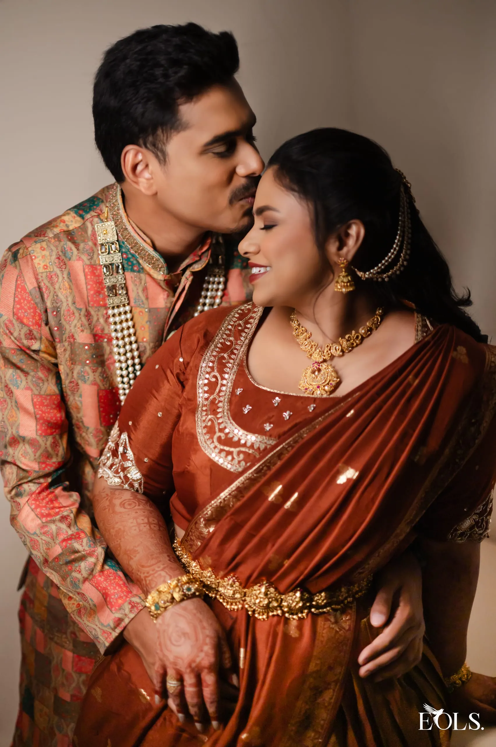 A man kisses a smiling woman’s forehead; both in traditional attire.
