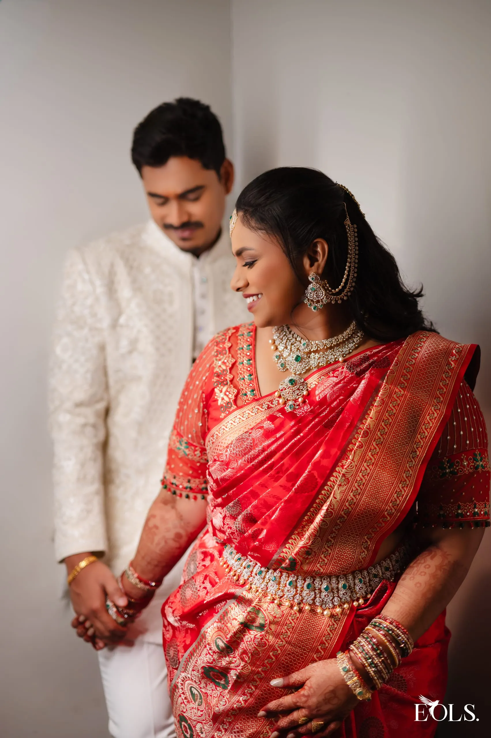 A woman in a red bridal saree and gold jewellery, a man behind her.