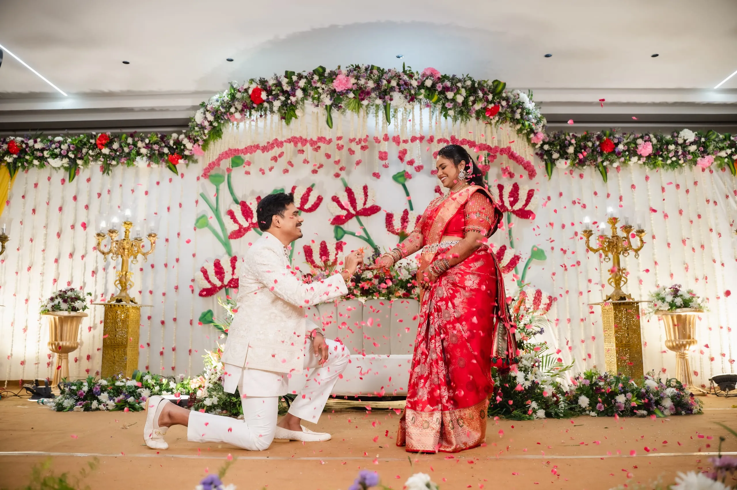 A man kneels to offer a ring on a flower-decked stage as petals fall.