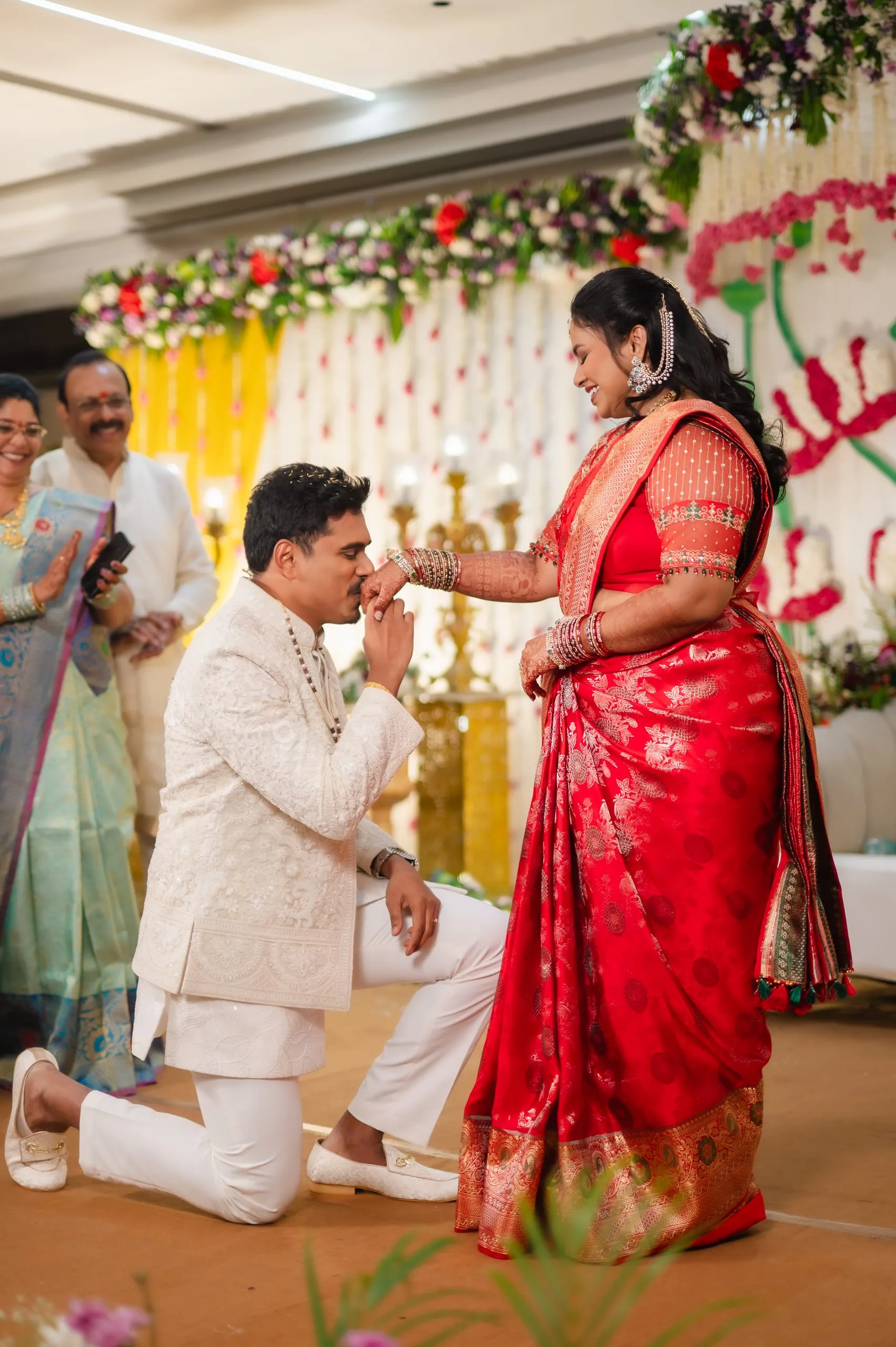 A kneeling man kisses a woman’s hand at a flowered stage.