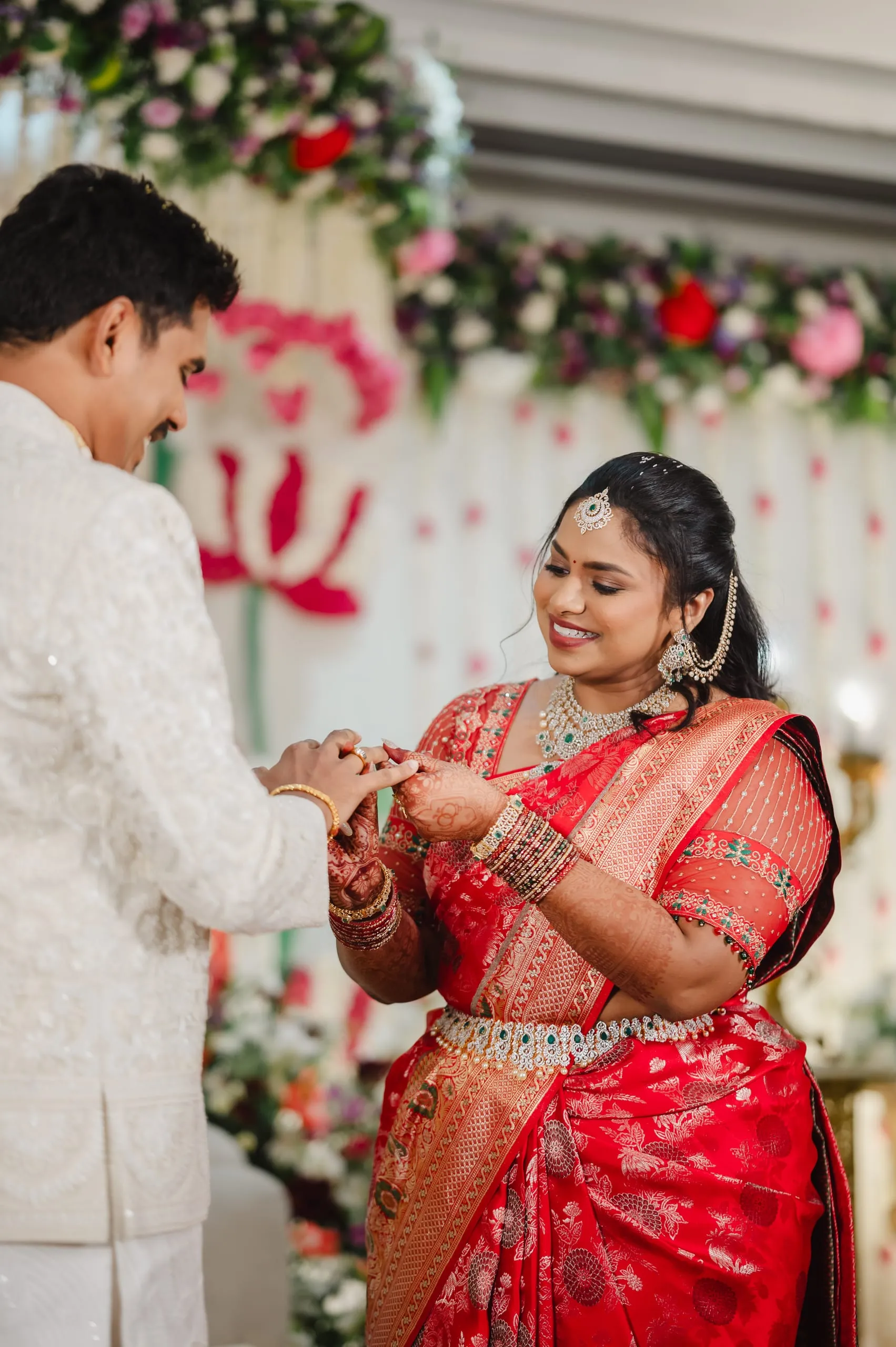 A woman places a ring on a man’s finger before a floral backdrop.