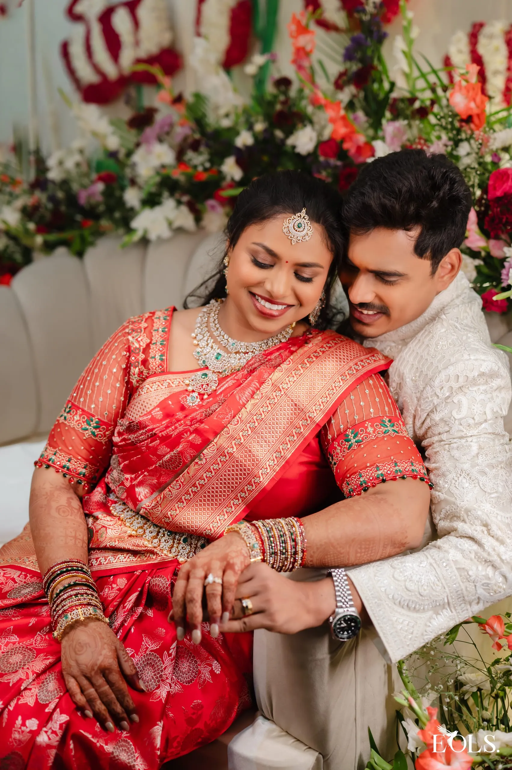 A couple sit close, eyes closed, before a large floral arrangement.
