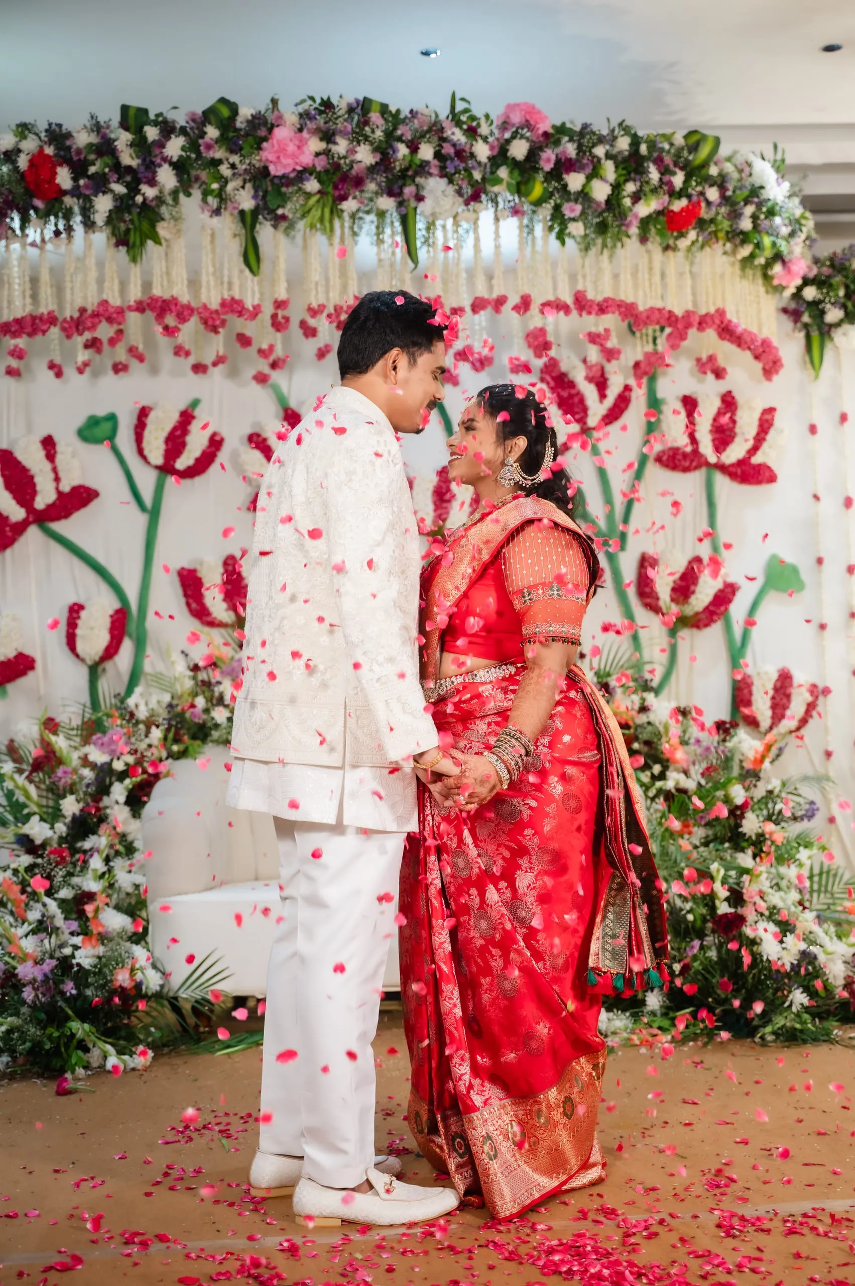 A couple hold hands amid falling petals before a floral backdrop.