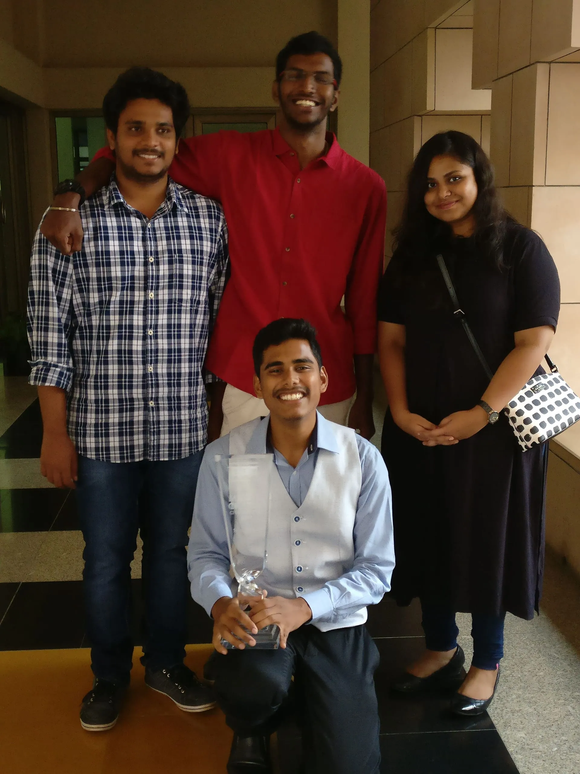 Four friends pose indoors, one holding a glass trophy.