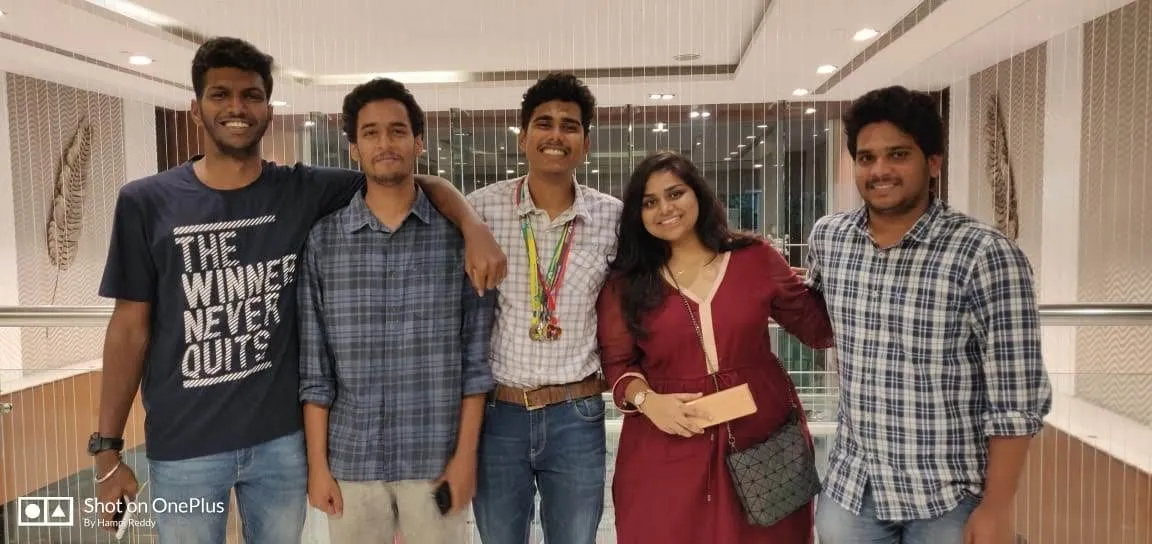 Five friends arm-in-arm indoors, one wearing medals.