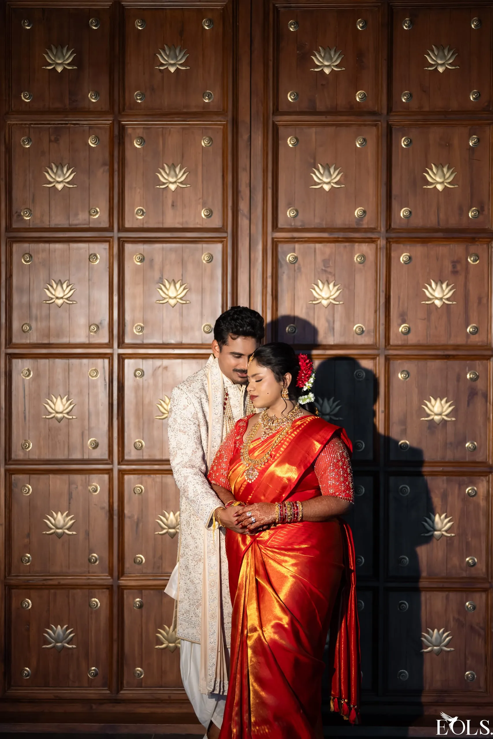 A groom embraces a bride from behind against a carved wooden door.