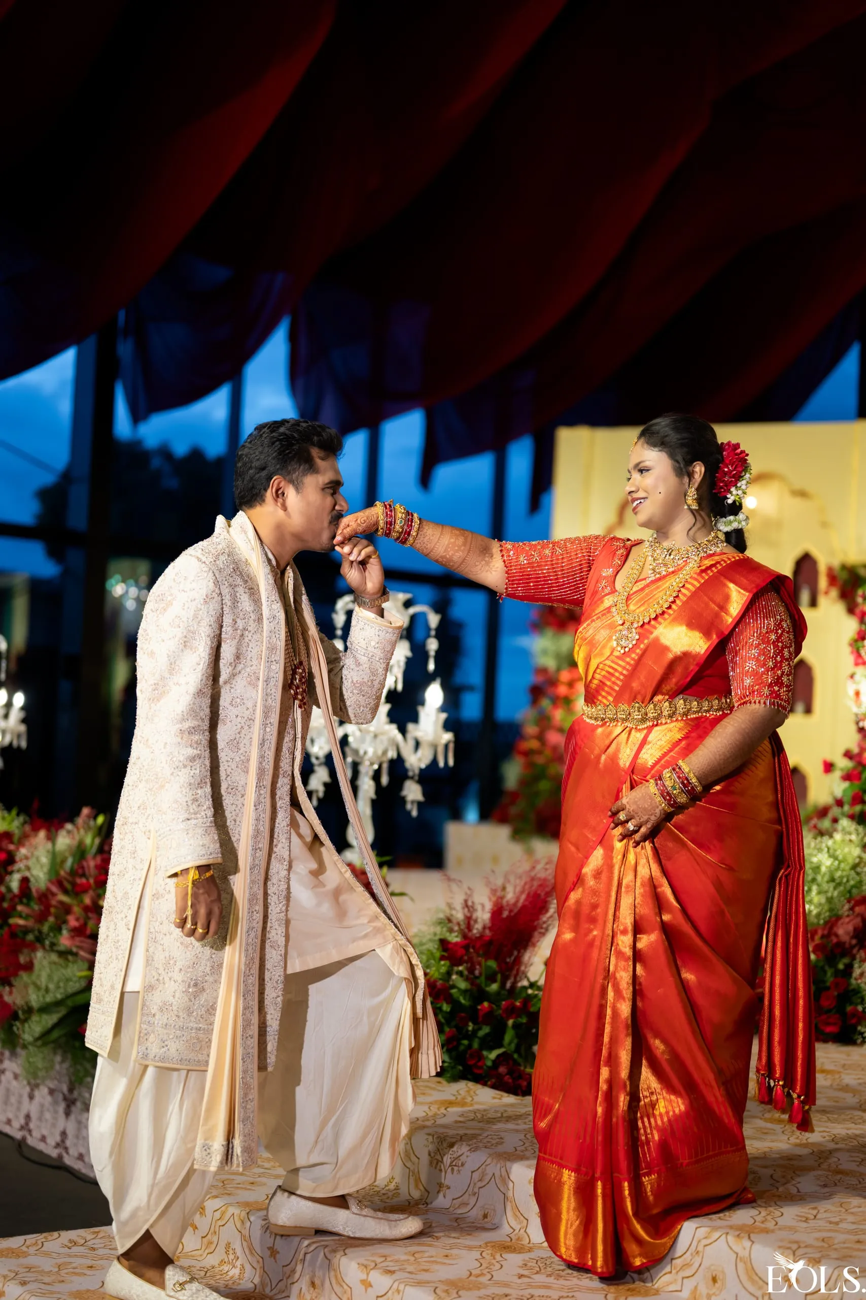 A groom kisses a bride’s hand on a floral stage at dusk.