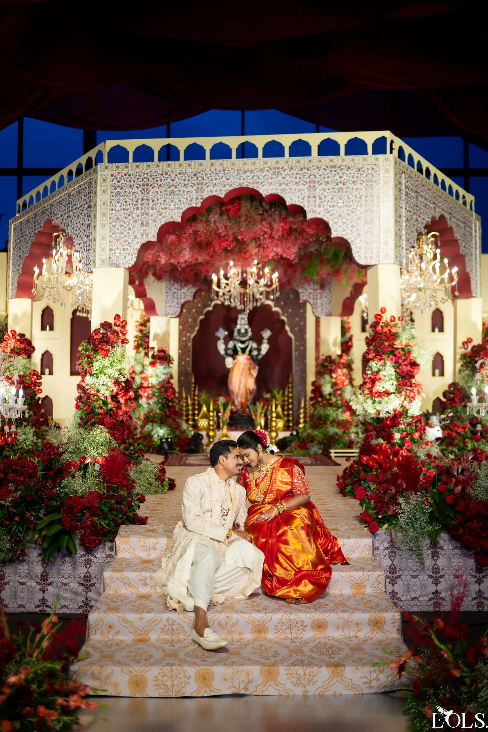 A couple seated on the steps of an ornate floral mandap.