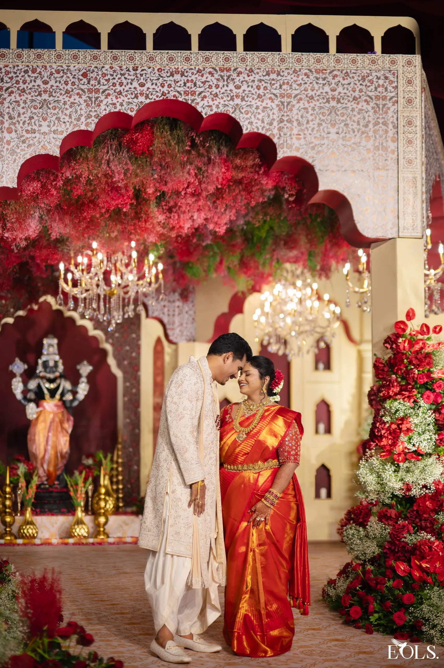 A couple forehead to forehead beneath a red floral arch.