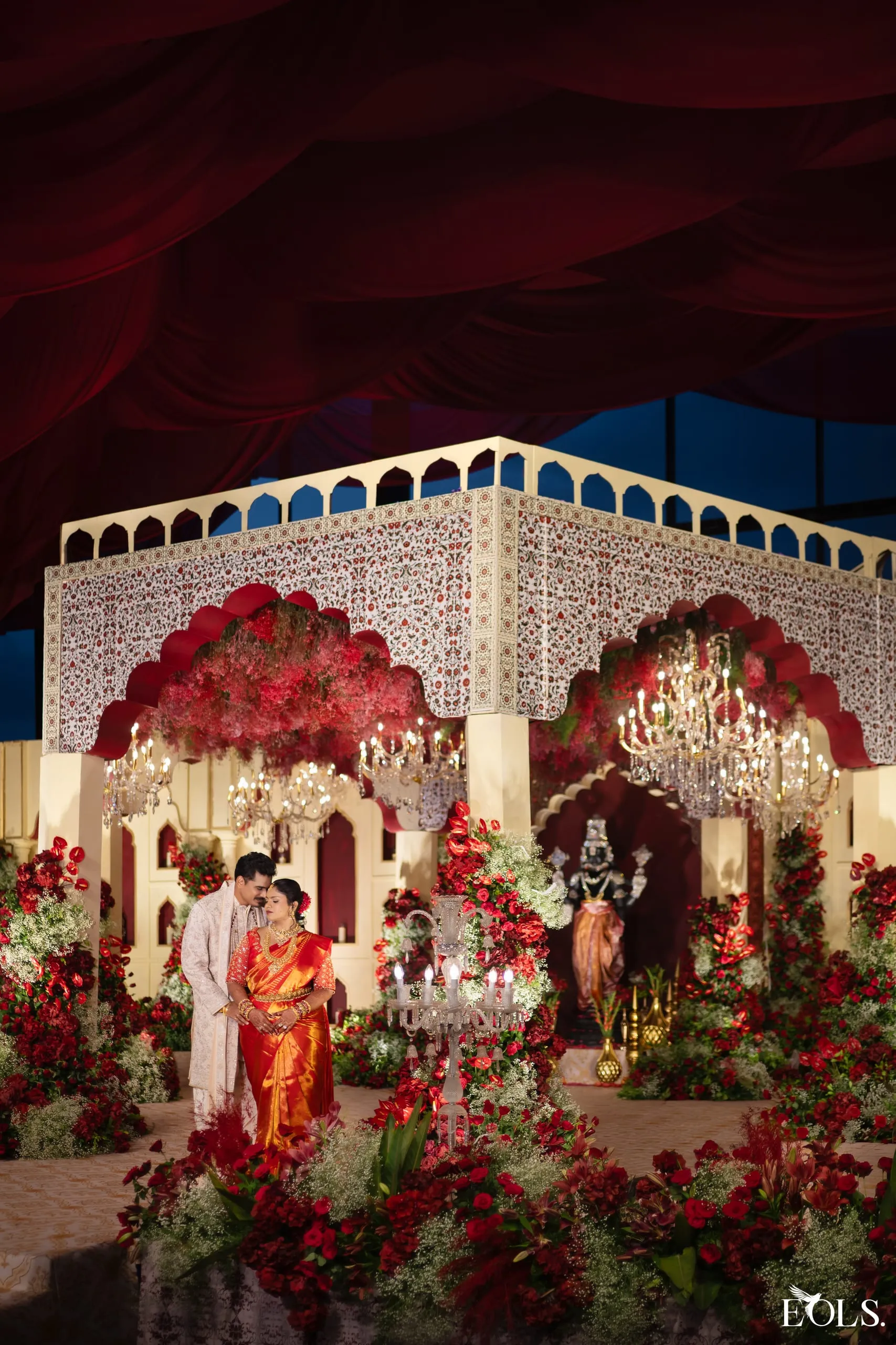 A couple in a grand mandap lavished with red roses and chandeliers.