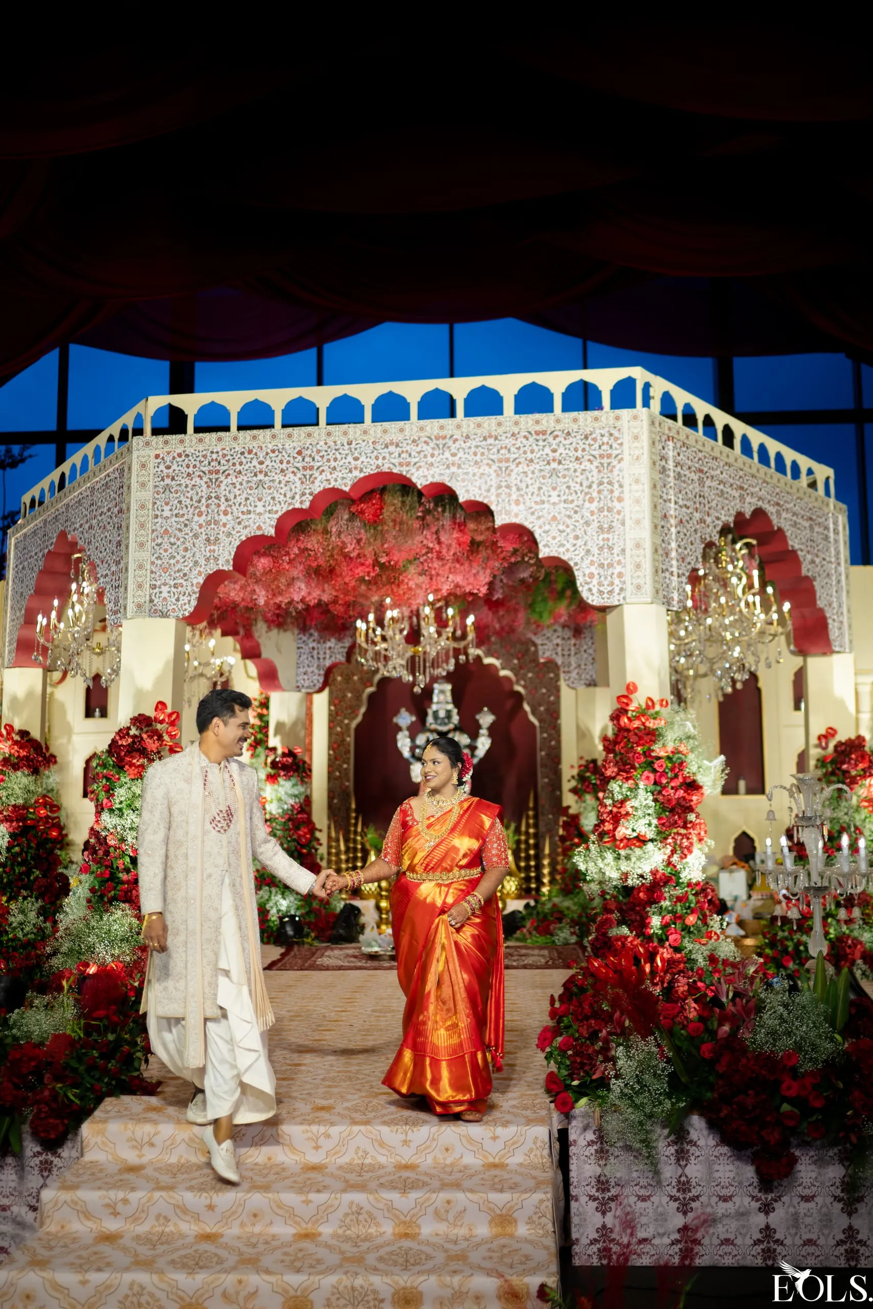 A couple walk down mandap steps hand in hand, smiling.